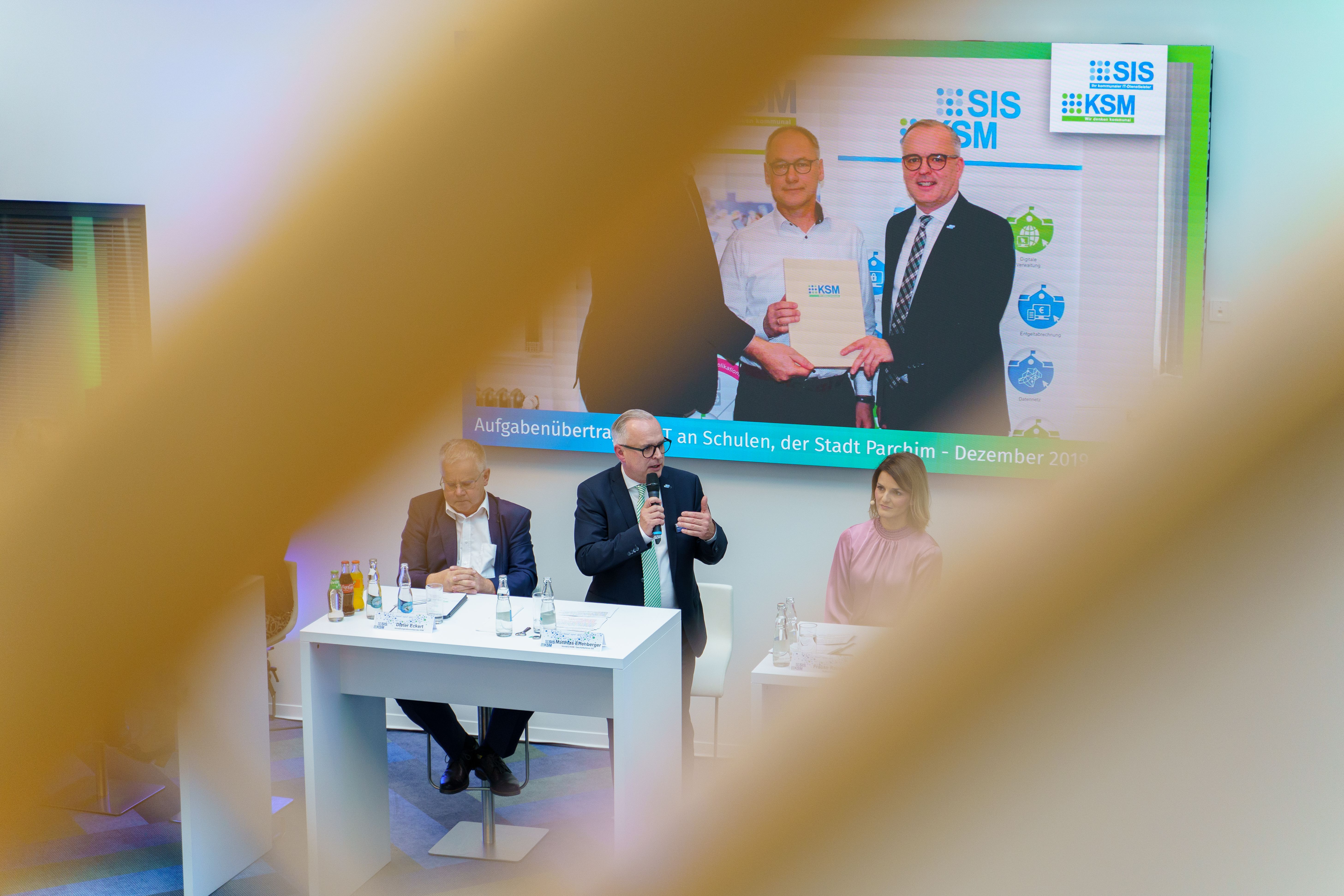 Redner mit Mikrofon am Podiumstisch, Leinwand im Hintergrund, Bokeh im Vordergrund