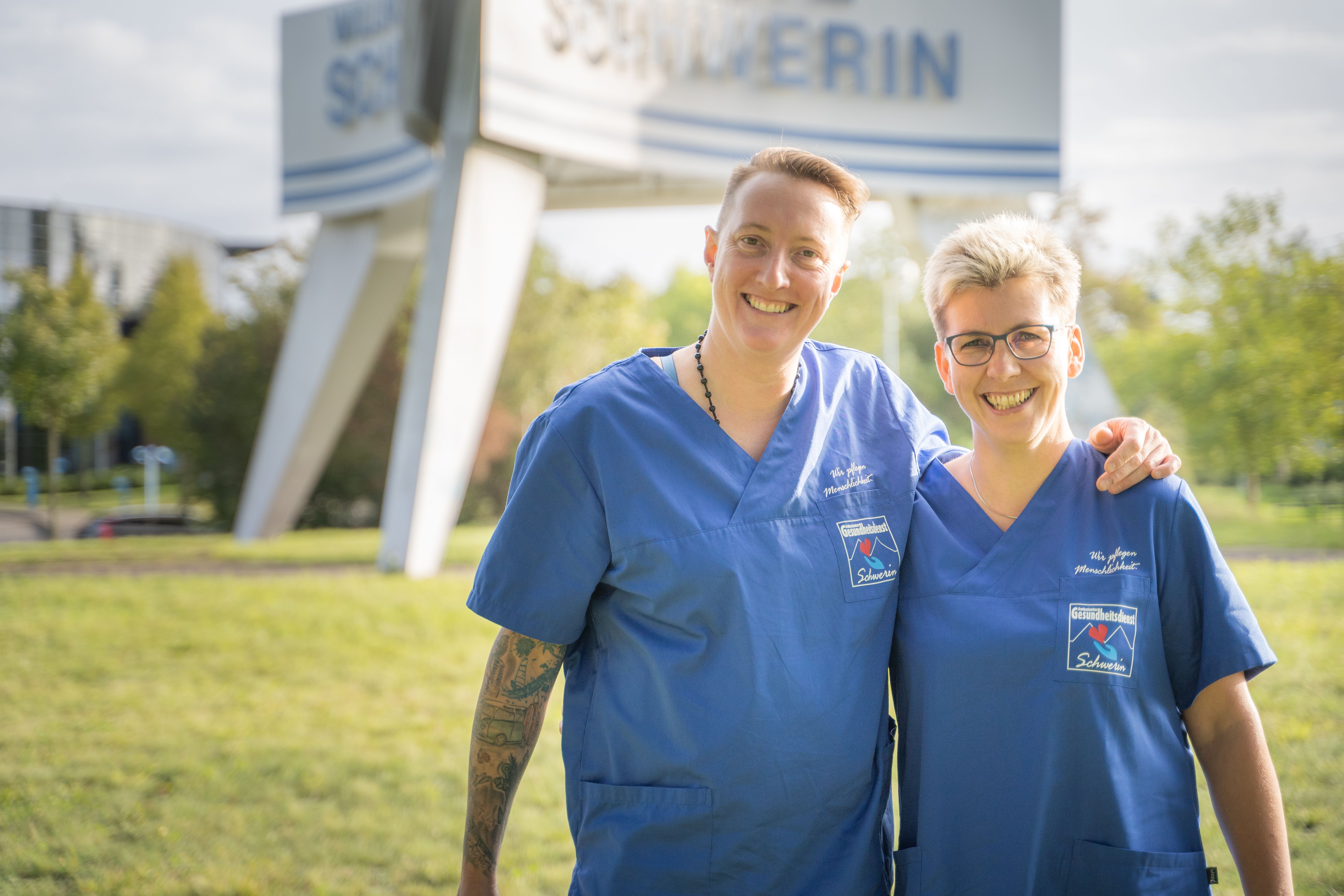 Zwei Frauen in blauer Berufskleidung umarmen sich lächelnd auf einer Wiese