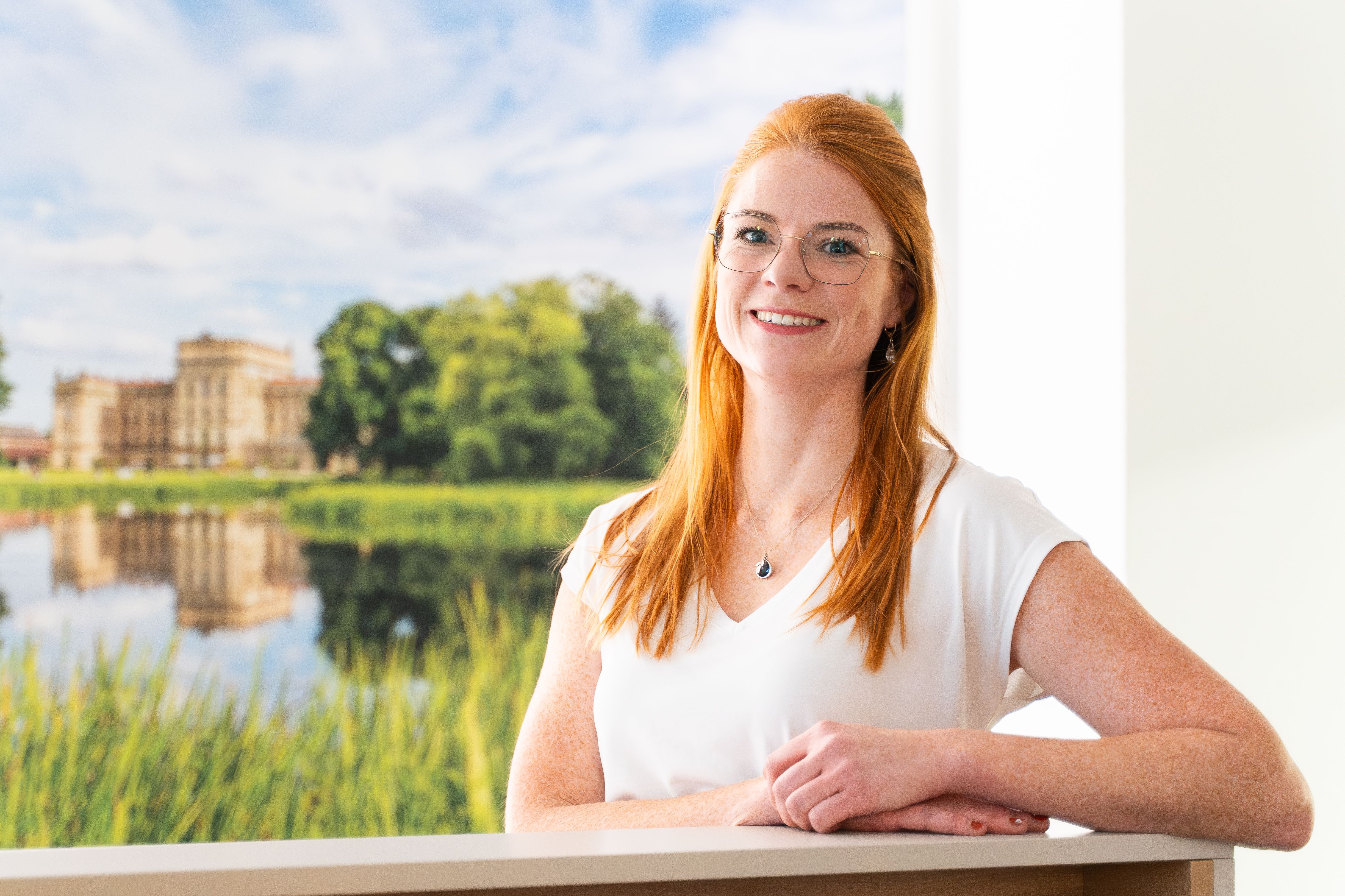 Lächelnde Frau mit roten Haaren vor einem Fenster mit Blick auf Schloss und See