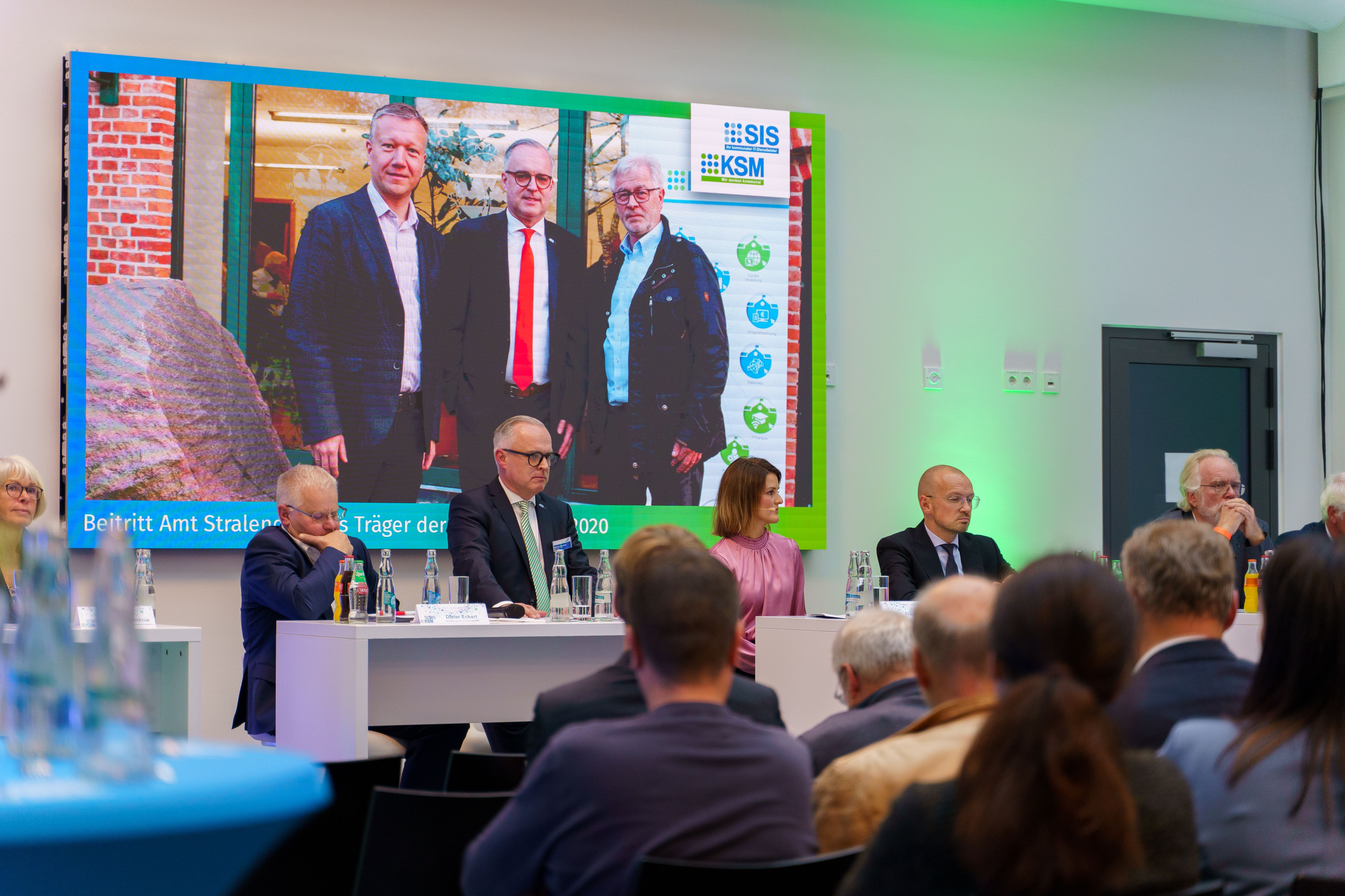 Podiumsdiskussion vor Publikum, Leinwand mit Gruppenfoto im Hintergrund