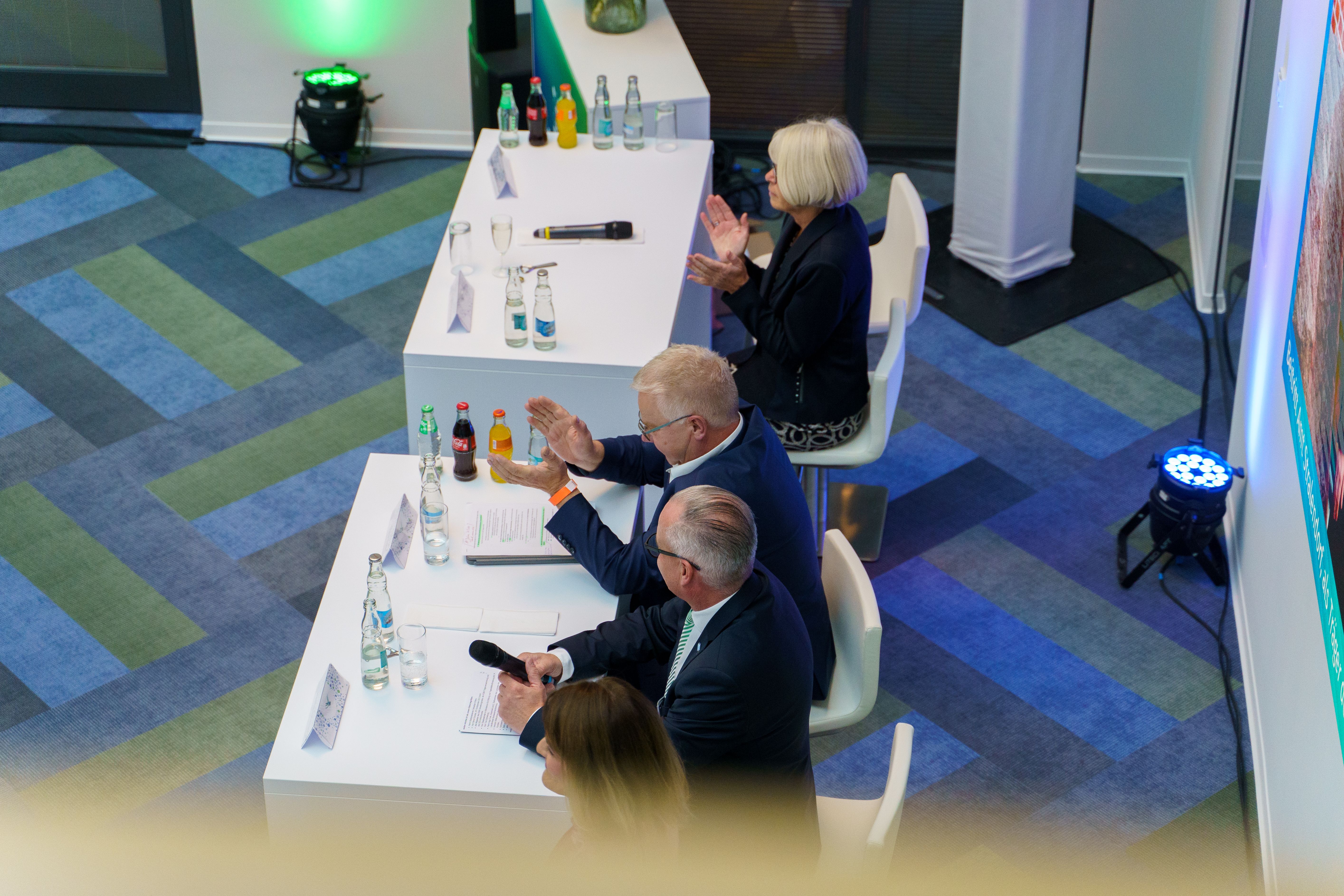 Podium bei einer Veranstaltung von oben, Menschen am Tisch applaudieren