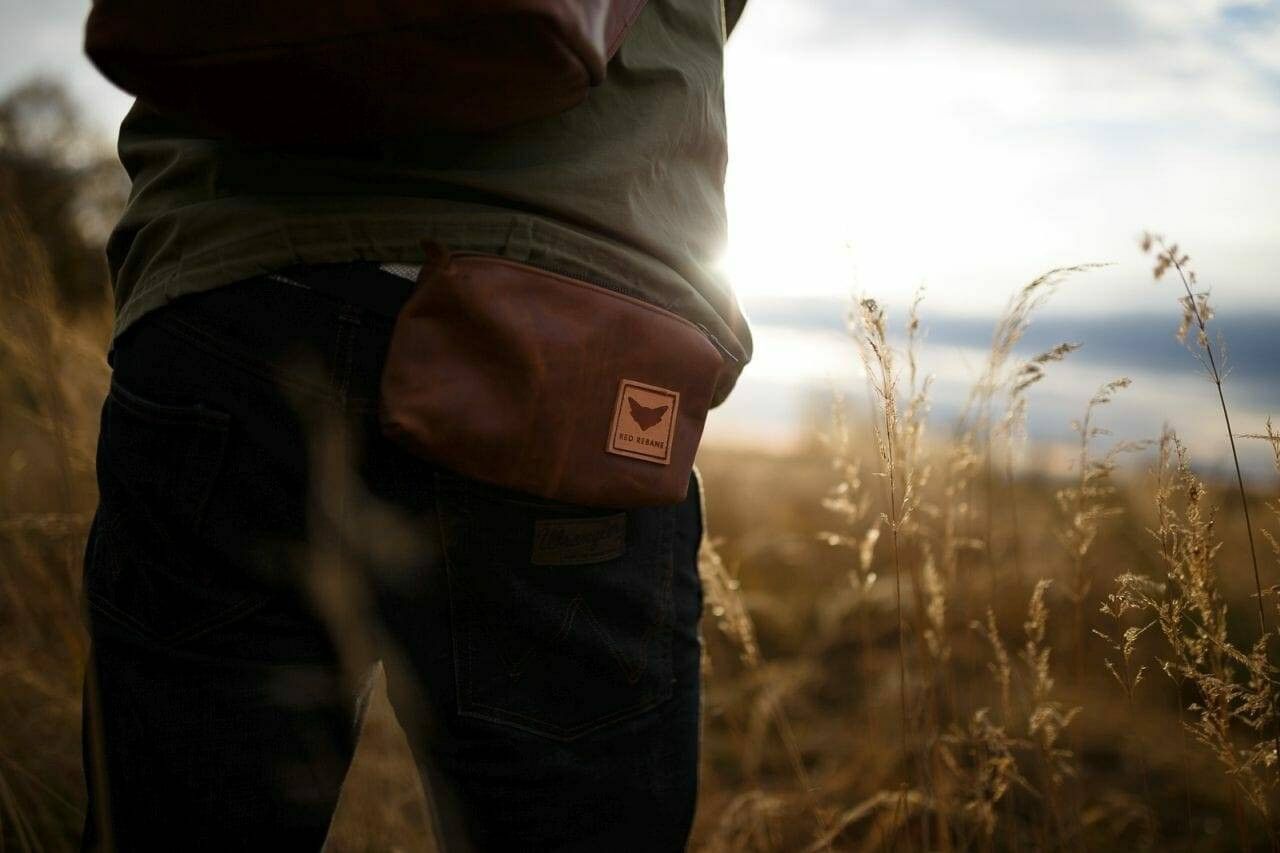 Bauchtasche aus braunem Leder an der Hüfte, dahinter Gräser im Gegenlicht
