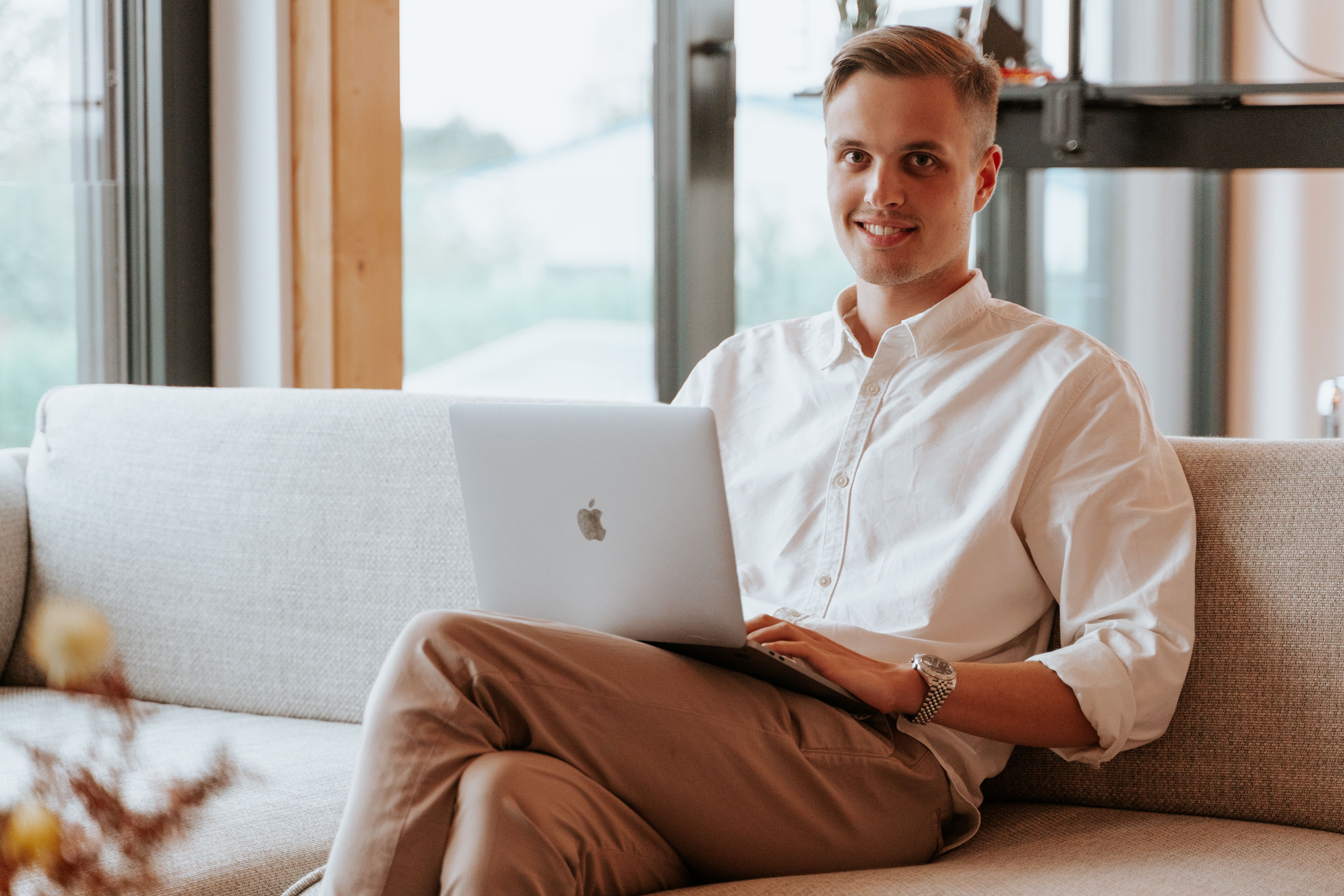Lächelnder junger Mann sitzt mit Laptop auf einem Sofa im hellen Büro