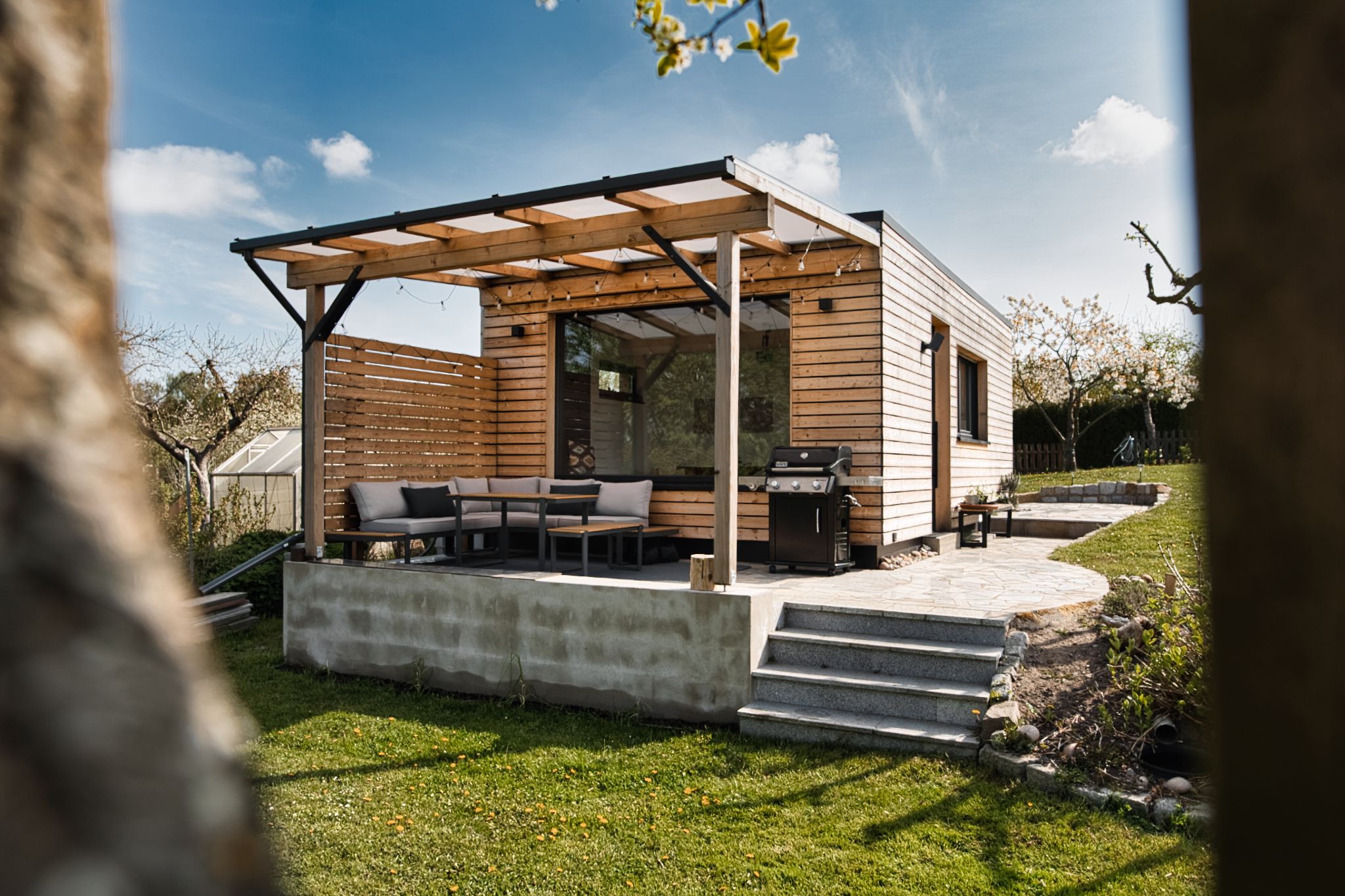 Gartenhaus aus Holz mit Terrasse und Sitzecke auf einer Wiese, blauer Himmel