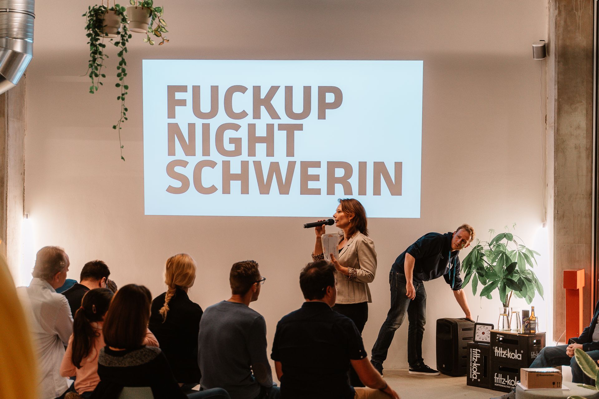 Rednerin mit Mikrofon vor sitzendem Publikum, Leinwand im Hintergrund