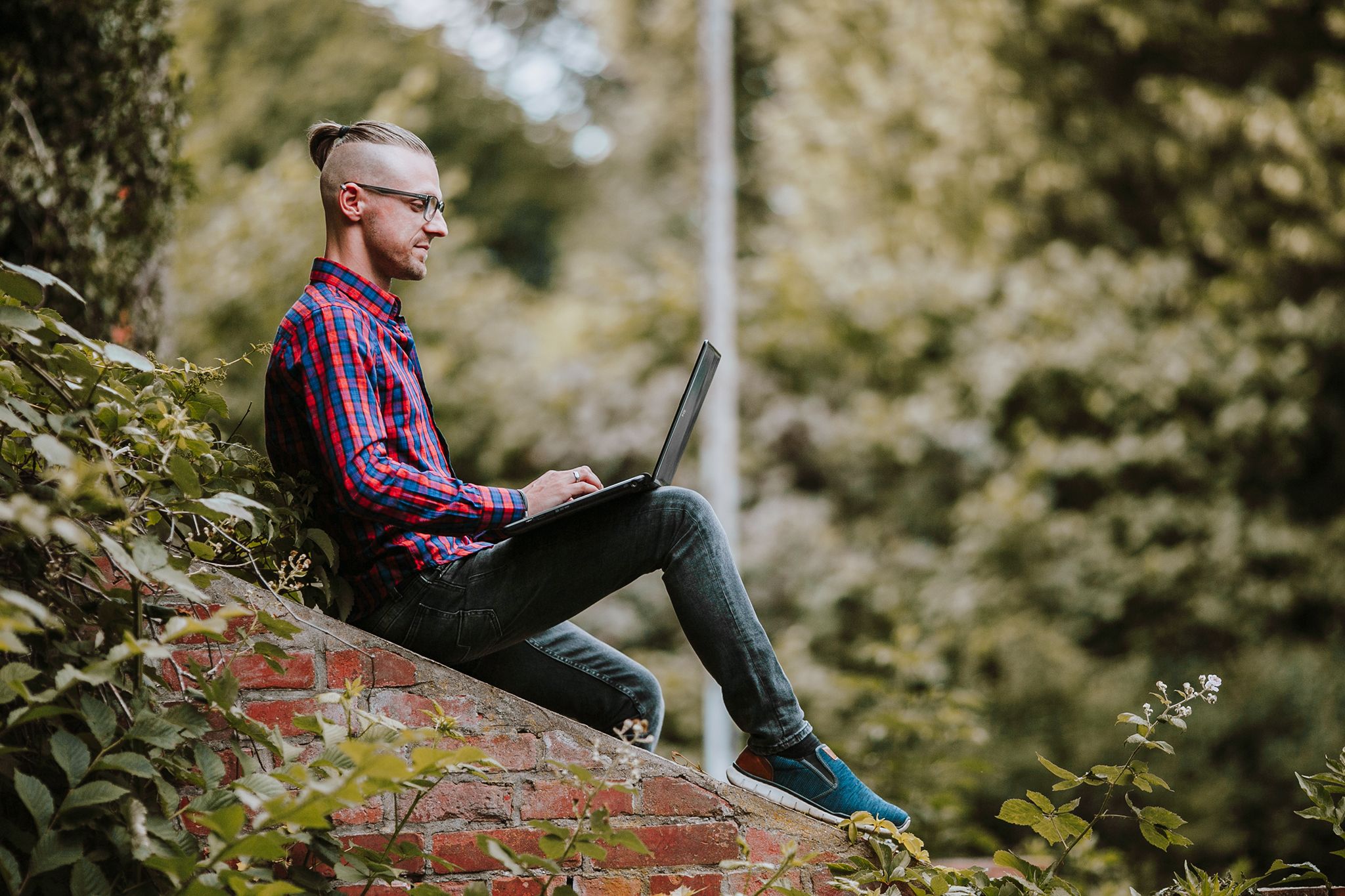 Mann sitzt mit Laptop auf einer Mauer im Grünen, weiches Licht