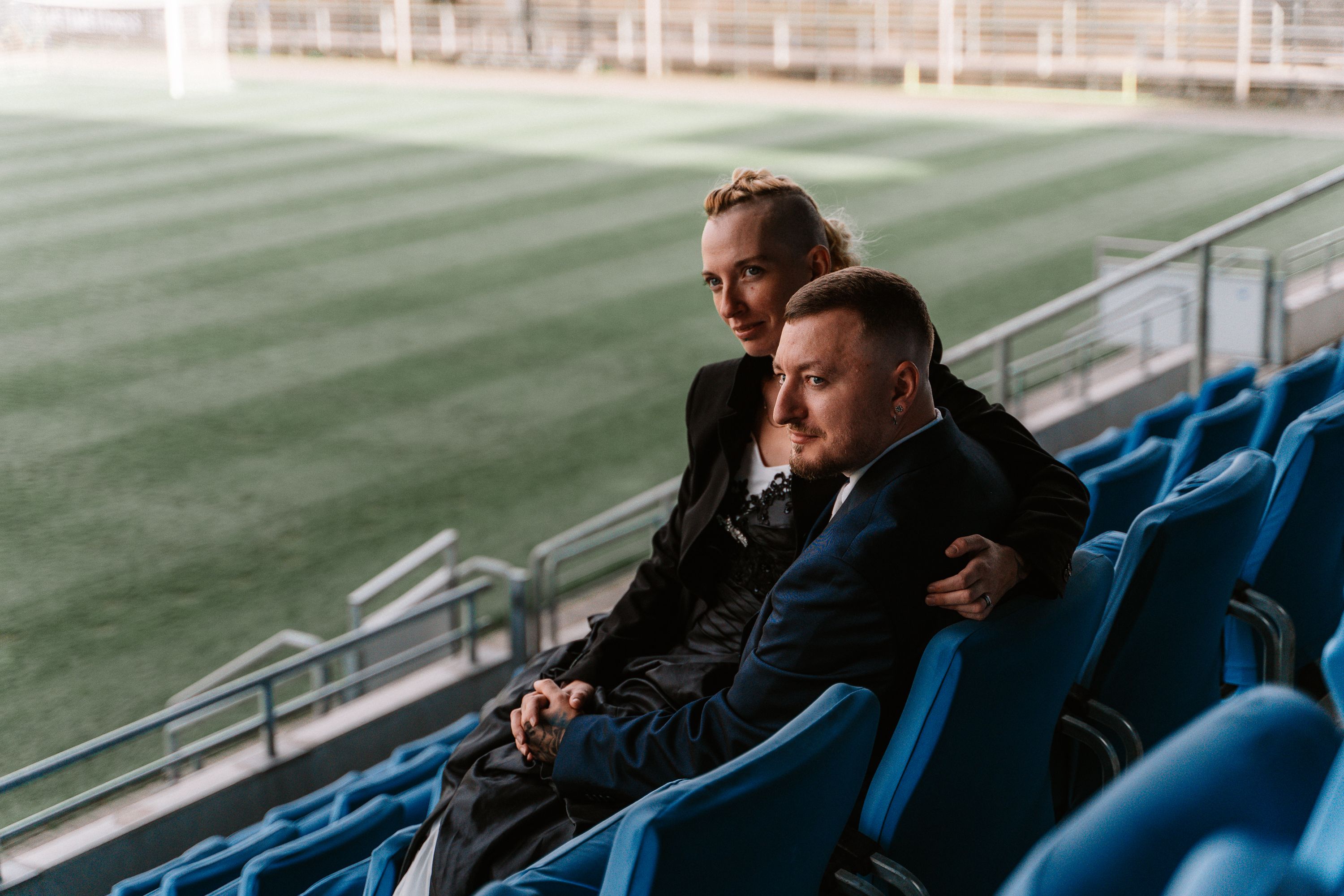 Zwei Männer in Hochzeitskleidung sitzen auf blauen Stadionsitzen vor dem Rasen