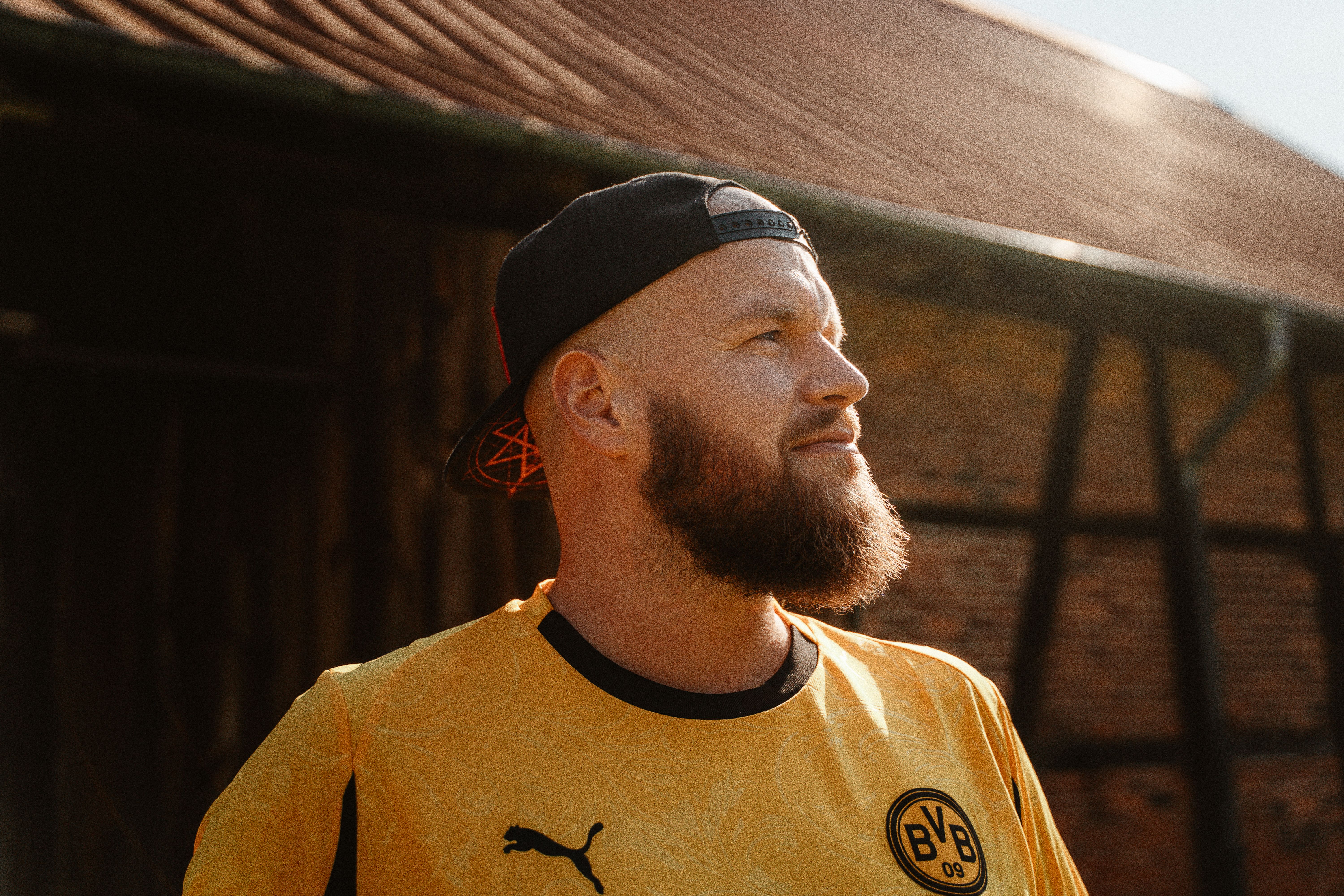 Mann mit Bart und Basecap im gelben Trikot vor einer Fachwerkscheune, Sonnenlicht